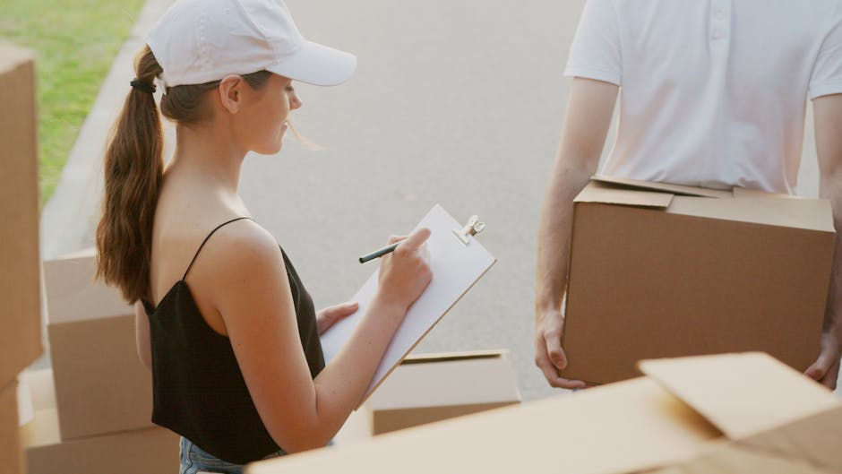 A woman with long hair tied in a ponytail, wearing a white cap and a black sleeveless top, is standing outdoors and writing on a clipboard with a pen. She is surrounded by several cardboard boxes, som