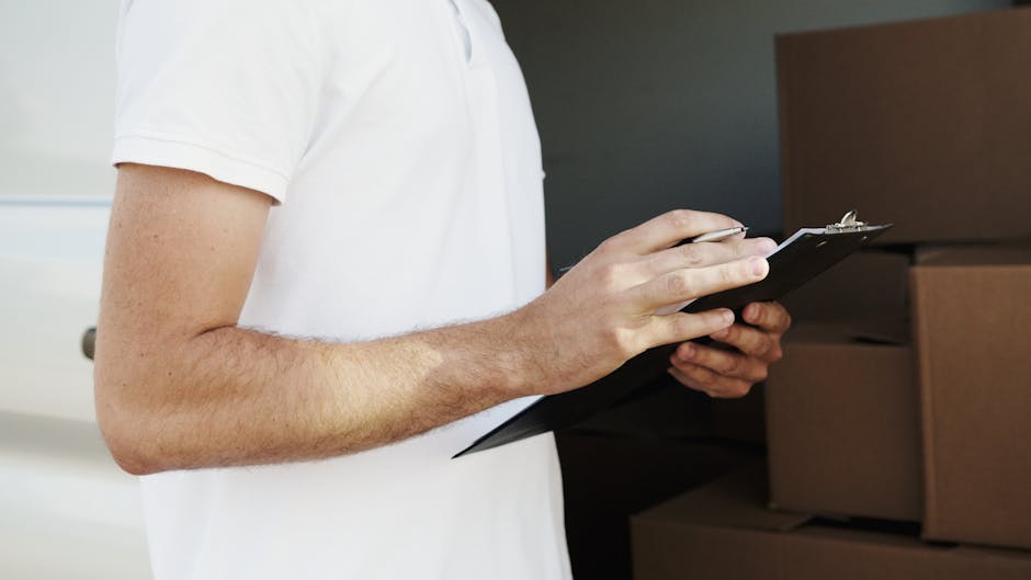 A woman with long hair tied in a ponytail, wearing a white cap and a black sleeveless top, is standing outdoors and writing on a clipboard with a pen. She is surrounded by several cardboard boxes, som