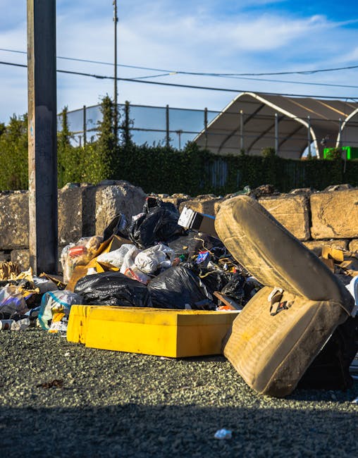 A pile of mixed bulky waste and household garbage, including large black and yellow plastic trash bags, a discarded old upholstered armchair with fabric upholstery, a yellow plastic storage container, and various cardboard and plastic packaging materials, all situated on a gravel surface near a stone wall and metal posts. In the background, there are power lines, trees, and a building with a curved roof under a partly cloudy sky. The scene appears to be outside a property, possibly during a home removal or clearance process, with no active loading or moving equipment visible. The presence of debris and waste materials reflects the clearing or disposal phase often associated with house removals or furniture transport services, as handled by companies like Man With a Van Northwood.