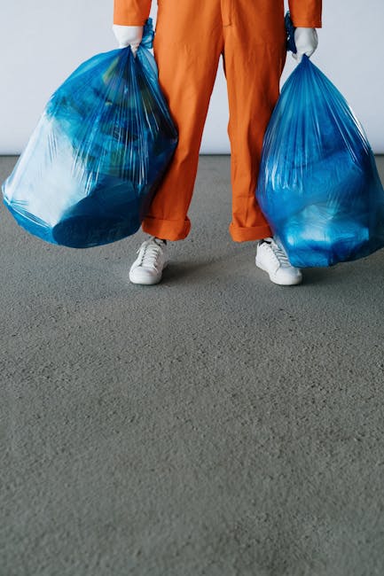 A person standing indoors on a grey carpeted floor is holding two large blue plastic garbage bags filled with household waste or recyclables, with their legs slightly apart. They are dressed in orange work trousers, a grey long-sleeved top, and white sneakers. The scene suggests a home cleaning or debris removal process as part of a house move or renovation. Behind the person's waist, a section of a white wall and a doorway leading to another room are visible. The image captures the practical aspect of packing or clearing out items during a household removal, with the usage of sturdy garbage bags indicating waste disposal or bulk removal tasks. This scene is relevant to the services offered by Man With a Van Northwood, particularly in relation to house removals, packing, and furniture transport in Northwood.
