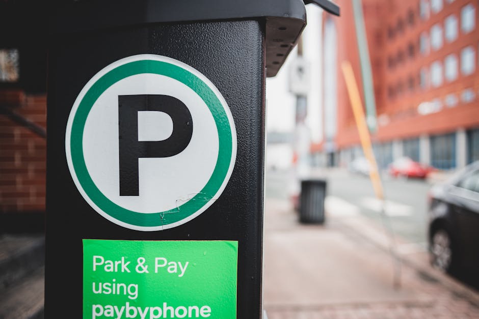 Close-up of a black parking meter on a city sidewalk, featuring a large white circular sign with a black 'P' indicating parking. Beneath the sign, a bright green sticker reads 'Park & Pay using paybyphone.' In the background, the street shows parked cars, red brick buildings, and a slightly blurred urban environment with a trash bin and building facades, suggesting a typical setting for home relocation or furniture transport services. The lighting is natural, and the focus is on the parking meter, which is relevant to vehicle access and moving logistics in Northwood as part of local removals arrangements performed by Man With a Van Northwood.