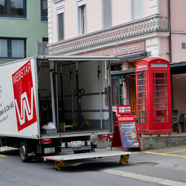 A white moving van with the logo 'WEBSTAR' on the side, parked on a city street outside a building with pastel green and pink facades, features an open rear loading door revealing empty interior space with a few tools and packaging materials inside, situated on a ramp with yellow and black safety stoppers. Next to the van, a red traditional telephone box is positioned on the pavement, with a small blackboard sign displaying prices and service information nearby, and a building with large windows, decorative railings, and signage indicating a pub and garden above the entrance. The scene depicts the setup process for home relocation or furniture transport as part of a house removals service, with natural lighting and urban surroundings.