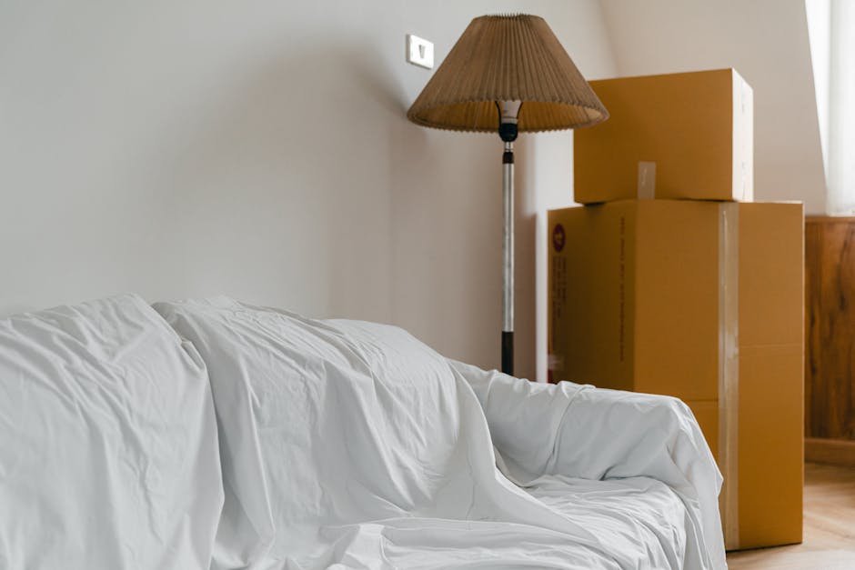 Interior view of a room showing a white upholstered sofa with visible fabric creases and a slightly wrinkled slipcover, positioned adjacent to a wall with a standard electrical socket mounted near the ceiling. To the right of the sofa, there are three brown cardboard moving boxes stacked on top of each other, with the largest box at the bottom and the smallest at the top, indicating a packing process associated with home relocation. Behind the boxes, a tall standing lamp with a beige fabric shade and a metal pole is visible, casting light onto the scene. The concrete or wooden flooring extends beneath the furniture and boxes, with natural light illuminating the space from a nearby window. The overall environment appears tidy, with furniture and packing materials prepared for removal or transport, aligning with professional house removals and moving services such as those offered by Man With a Van Northwood during emergency same-day relocations in Northwood.