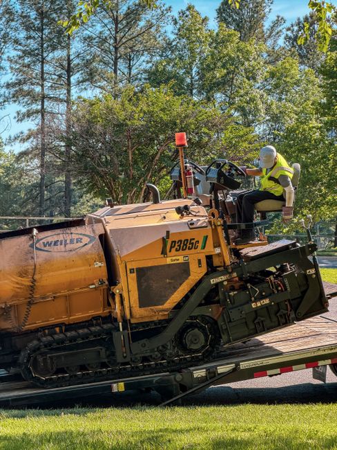A worker wearing a high-visibility yellow vest and a white helmet is seated in the driver’s seat of a large yellow and black Weiler P3850C industrial paving machine, which is being loaded onto a flatbed truck outside a residential property. The machine, used in driveway or road work, features a control panel with levers and a steering wheel. The flatbed truck, partially visible, is situated on a pavement adjacent to a well-maintained lawn. In the background, tall trees and green foliage suggest a suburban environment, with ample natural daylight illuminating the scene. This image captures the process of loading heavy equipment, typical in home relocation or building projects, supported by companies like Man With a Van Northwood, specialists in removals and moving logistics.