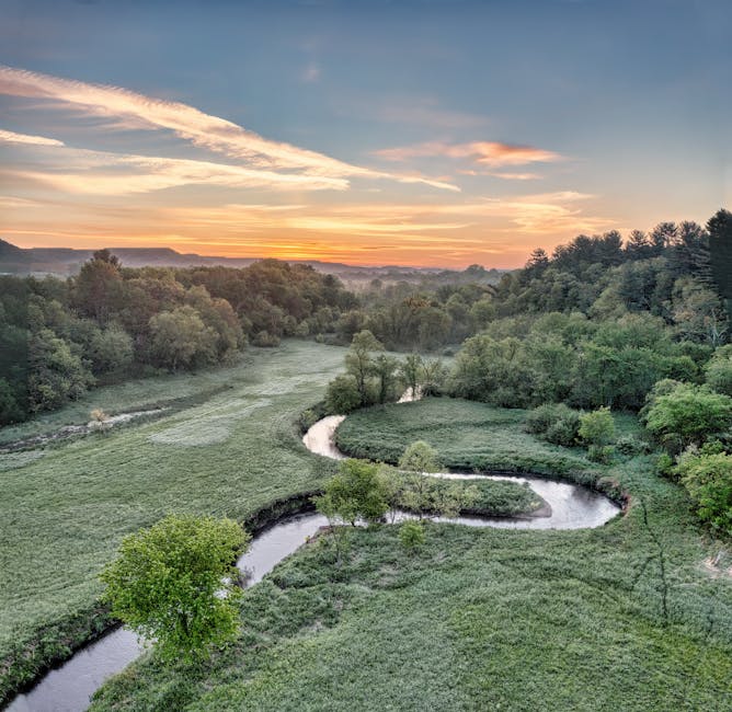Aerial view of a winding river flowing through lush green fields and dense woodland under a partly cloudy sky at sunset. The landscape features grassy meadows with scattered trees along the riverbanks, leading towards distant rolling hills. The scene is illuminated by warm, orange hues from the setting sun, casting gentle light on the natural environment. This tranquil rural scene exemplifies peaceful countryside scenery, with no signs of human activity or structures visible. The image captures the serene beauty of nature, suitable for a background or illustrative purpose related to home relocation or outdoor environments, as part of content for Man With a Van Northwood's removals and moving services.