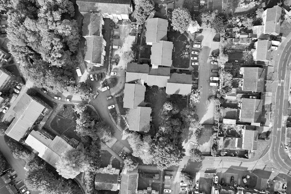 Aerial black and white photograph of a residential area showing multiple houses with pitched roofs, surrounded by trees and narrow roads with parked cars. In the foreground, a moving van is parked on a driveway, partially visible, with several cardboard boxes, wrapped furniture, and packing materials such as plastic wrap laid out nearby. A person is seen lifting or carrying a piece of furniture onto the van or from the driveway, indicating the process of home relocation or furniture transport. The scene includes tools like trolleys and straps visible on the ground, emphasizing loading activities. The environment appears in natural daylight, capturing the spatial arrangement of homes, vehicles, and moving equipment. The image, associated with Man With a Van Northwood and house removals services, reflects aspects of packing, loading, and transport logistics involved in house moves, supporting the context of moving regulations and procedures outlined by Hillingdon Council for Northwood property relocations.