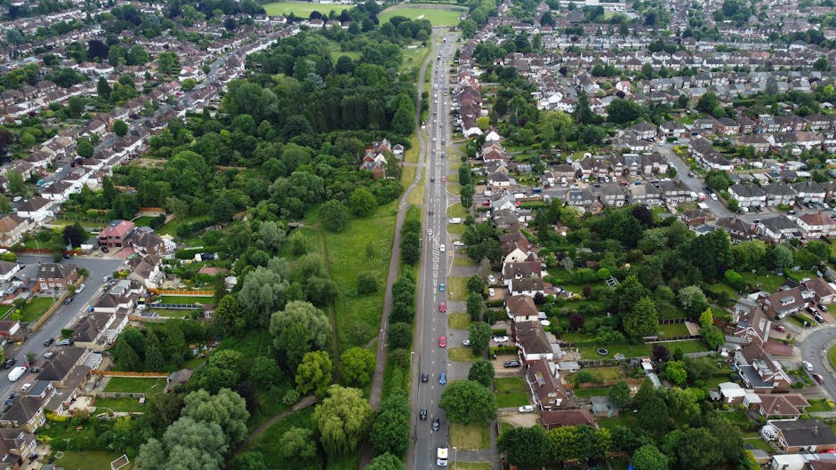 An aerial view of a residential neighbourhood showing a main road running through the centre, with parked cars along the kerbside and a line of houses on either side. To the left of the road, there is a large area of greenery consisting of trees, shrubs, and grass, with some pathways weaving through. On the right side, rows of terraced houses with driveways and small front gardens are visible, many with gardens and patio furniture. The street appears calm, with a few vehicles in motion. The scene is captured in daylight with clear visibility of the suburban environment. This image can support content about home relocations and moving logistics, highlighting the environment where Man With a Van Northwood might undertake property removals and furniture transport services, particularly when planning a move or packing process in Northwood.