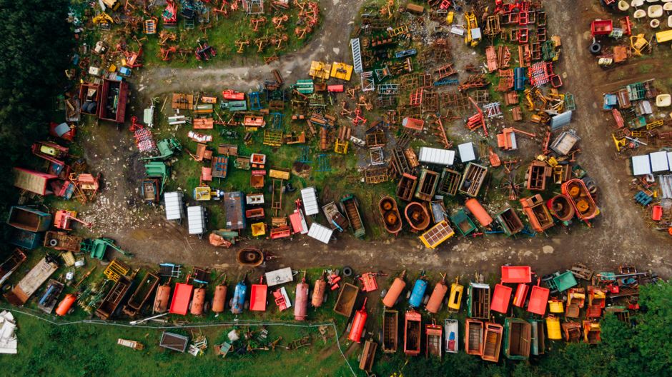 An aerial view of a large outdoor area filled with various discarded items and farm machinery, including tractors, trailers, and metal equipment in multiple colors such as red, green, yellow, and orange. The equipment is arranged in disorganized clusters across patches of grass and dirt pathways, suggesting a scrapyard or storage yard for agricultural tools and vehicles. The lighting appears natural, with some shadows indicating daylight. The scene reflects a cluttered outdoor space where machinery is likely being prepared for disposal or transfer. Based in Northwood, Man With a Van Northwood specializes in house removals and furniture transport, supporting efficient logistics during home relocation or moving processes, often involving careful handling of equipment and tools like those visible in the image.