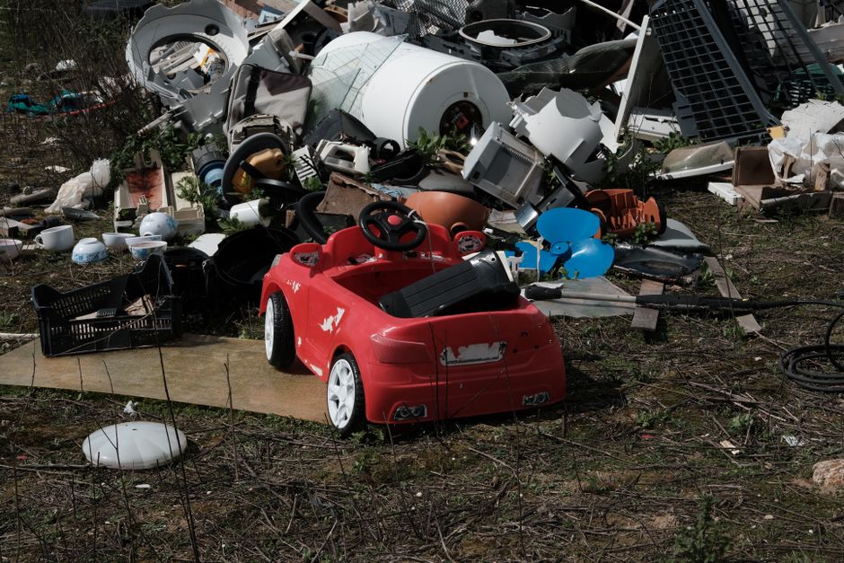 An outdoor scene showing a large pile of discarded household items and rubbish, including broken appliances, plastic containers, and miscellaneous debris, with a small red battery-operated toy car positioned on a flat wooden board in the foreground. The toy car, designed to resemble a compact vehicle, has black seats and a black steering wheel, and is placed amid the debris, which is spread across a patch of bare ground with some grass and weeds. The background features additional clutter such as inflatable items, plastic baskets, and various broken furnishings, all amidst a wire fence. The scene appears to depict waste or waste collection, possibly related to junk removal or disposal services by Man With a Van Northwood, relevant to house removals and clearance tasks, with natural lighting highlighting the details of the clutter and scavenged items.