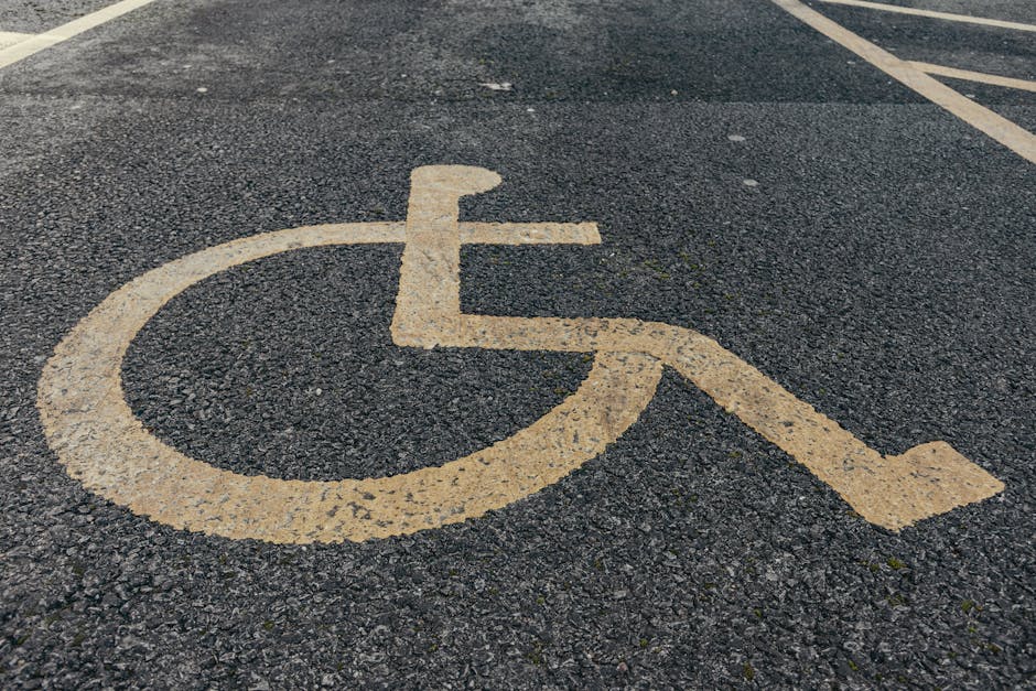 A close-up view of a weathered yellow wheelchair-accessible parking symbol painted on a dark asphalt surface outside a property, with visible cracks and texture in the pavement. The painted symbol features a stylised wheelchair icon with a circular wheel at the front and a curved backrest, accompanied by directional lines indicating parking space boundaries. In the background, parts of another parking space outline can be seen, along with faint white lines marking individual spots. The scene suggests an outdoor parking area associated with a residential or commercial premises, likely used for facilitating home relocations or furniture transport for clients of Man With a Van Northwood, as seen on their webpage about access and parking solutions for house removals in Northwood.