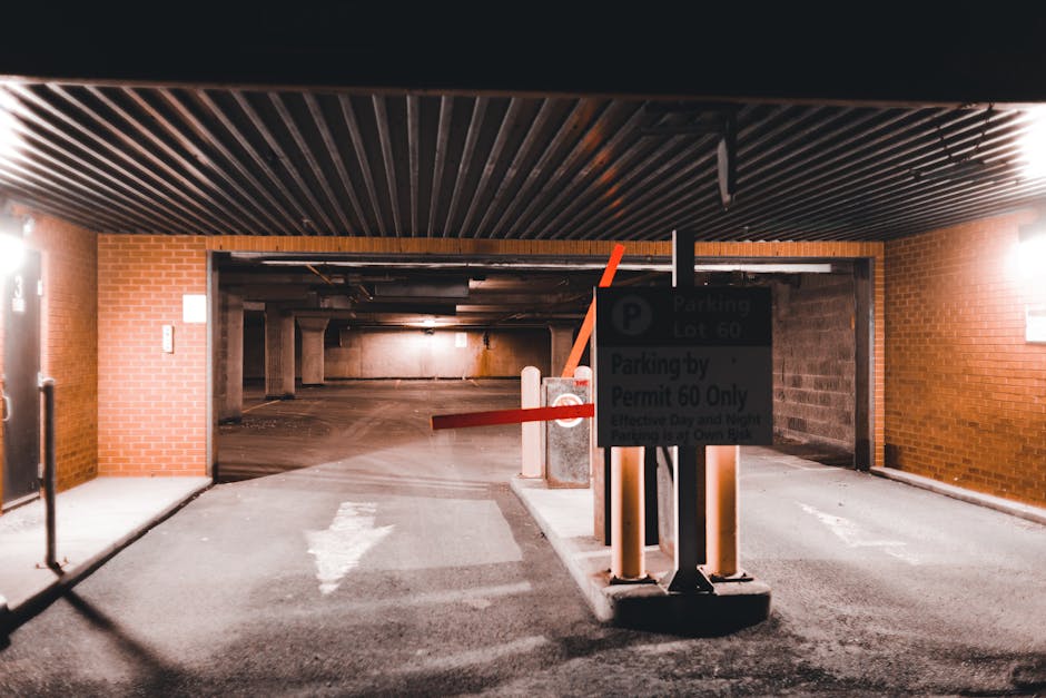The image shows the entrance to an underground parking garage with a controlled access barrier arm in a horizontal position, preventing vehicle entry. In the foreground, the barrier arm is supported by three concrete pillars, and a sign displaying parking restrictions and permit information is mounted on a metal post nearby. The ground is paved with asphalt and features a white painted arrow indicating the direction of traffic flow into the parking area. To the left, there is a brick wall with a lamp providing illumination, and a metal gate with a lock is visible further along the wall. Inside the garage, the ceiling consists of dark, horizontal wooden slats, and the lighting is dim with a reddish glow, suggesting artificial illumination. The background reveals a spacious, multi-level parking area with concrete columns and a slightly curved floor, suitable for vehicle movement and parking coordination, aligning with house removal and moving logistics managed by Man With a Van Northwood for projects such as home relocation and furniture transport.