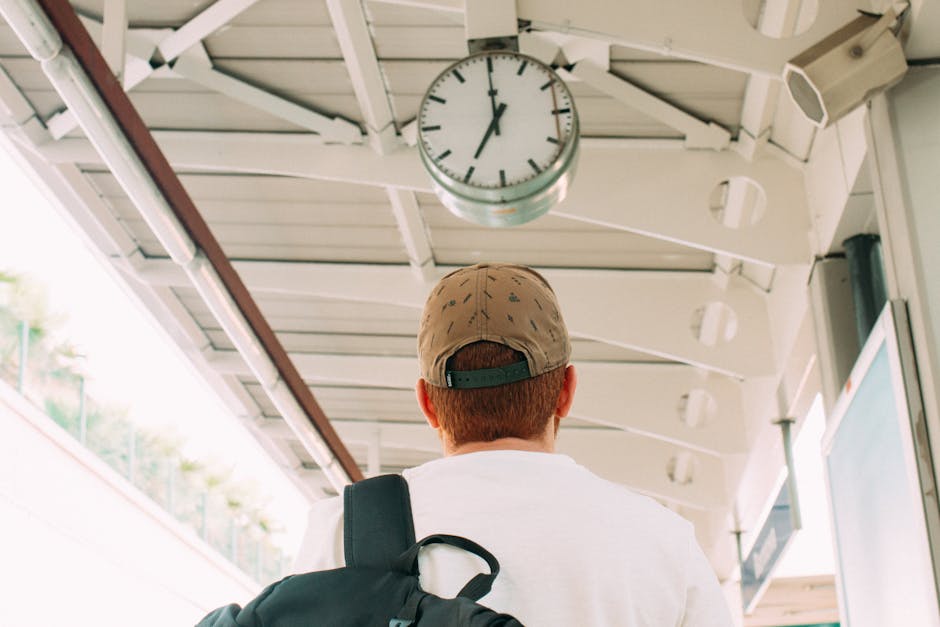 A man with red hair, wearing a beige camo baseball cap and a white shirt, stands at an outdoor train or bus station, facing away from the camera. He carries a black backpack over his right shoulder, and is looking up at a large round clock mounted on a metal beam ceiling. The station's roof structure is visible, made of white painted steel beams and panels, with small white lights attached along the ceiling edge. The background shows a bright day with green foliage and part of a platform or staircase. The scene captures a moment of waiting or planning during a home relocation, with the man possibly checking the time before a move or transport service provided by Man With a Van Northwood, as indicated by the context, with emphasis on the environment and moving logistics.