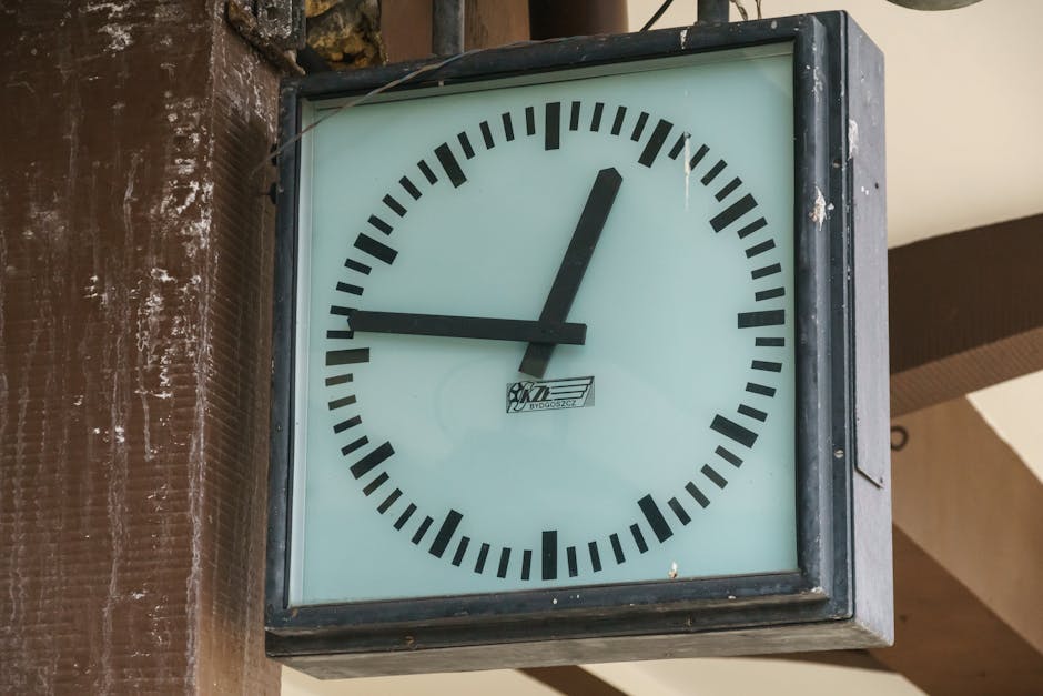A large, square outdoor wall clock mounted on a brown brick wall near a building structure. The clock face has a white background with black hour and minute hands; the hour hand points slightly past 12, and the minute hand points to 3, indicating approximately 12:15. The clock features black tick marks for each minute and bold black hour markers. The frame around the clock is metal, showing some signs of weathering. The lighting suggests daylight, with shadows cast by nearby structures. This clock is positioned at an accessible height, likely near a public space or station, supporting timing and scheduling during a home relocation or furniture transport process. The placement and visibility of the clock are relevant for coordinating loading and unloading moments during house removals, as provided by Man With a Van Northwood.