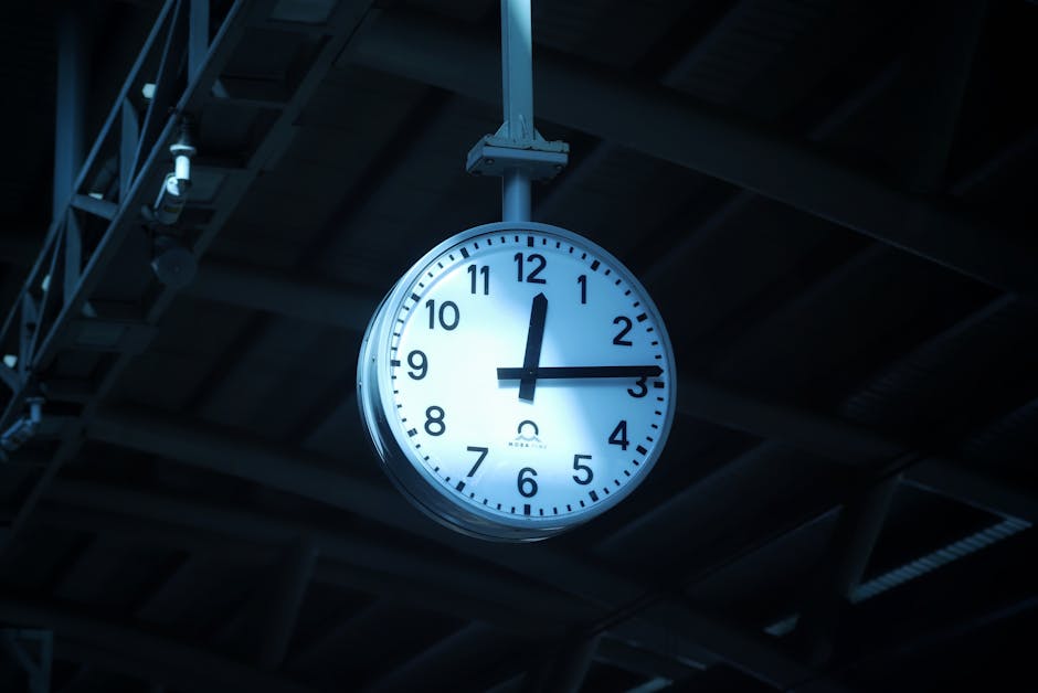 A man with red hair, wearing a beige camo baseball cap and a white shirt, stands at an outdoor train or bus station, facing away from the camera. He carries a black backpack over his right shoulder, and is looking up at a large round clock mounted on a metal beam ceiling. The station's roof structure is visible, made of white painted steel beams and panels, with small white lights attached along the ceiling edge. The background shows a bright day with green foliage and part of a platform or staircase. The scene captures a moment of waiting or planning during a home relocation, with the man possibly checking the time before a move or transport service provided by Man With a Van Northwood, as indicated by the context, with emphasis on the environment and moving logistics.