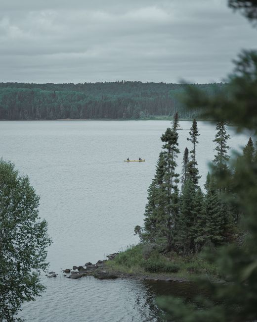 A tranquil lakeside scene showing a calm body of water surrounded by lush green trees with a dense forest in the background. In the middle of the lake, a small yellow boat with two occupants is visible, possibly engaged in fishing or leisure activity. The shoreline features a few rocks and vegetation, with an overcast sky casting diffuse light over the landscape. Though the image does not depict house removals, it portrays a peaceful environment that could relate to outdoor relocation or nature-based moving services. The natural scenery emphasizes quiet, scenic surroundings typical of Northwood and nearby areas where home relocation or outdoor packing and transport might take place, relevant to services offered by companies like Man With a Van Northwood.